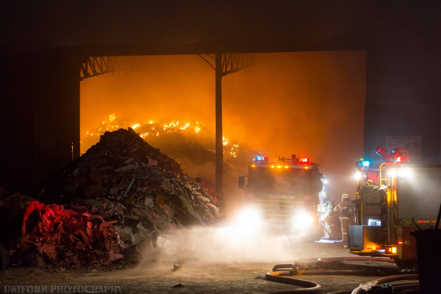 The picture shows a large pile of waste is alight in the background and two FRV fire appliances and two firefighters wearing breathing apparatus at the side of the truck adjusting the pump.