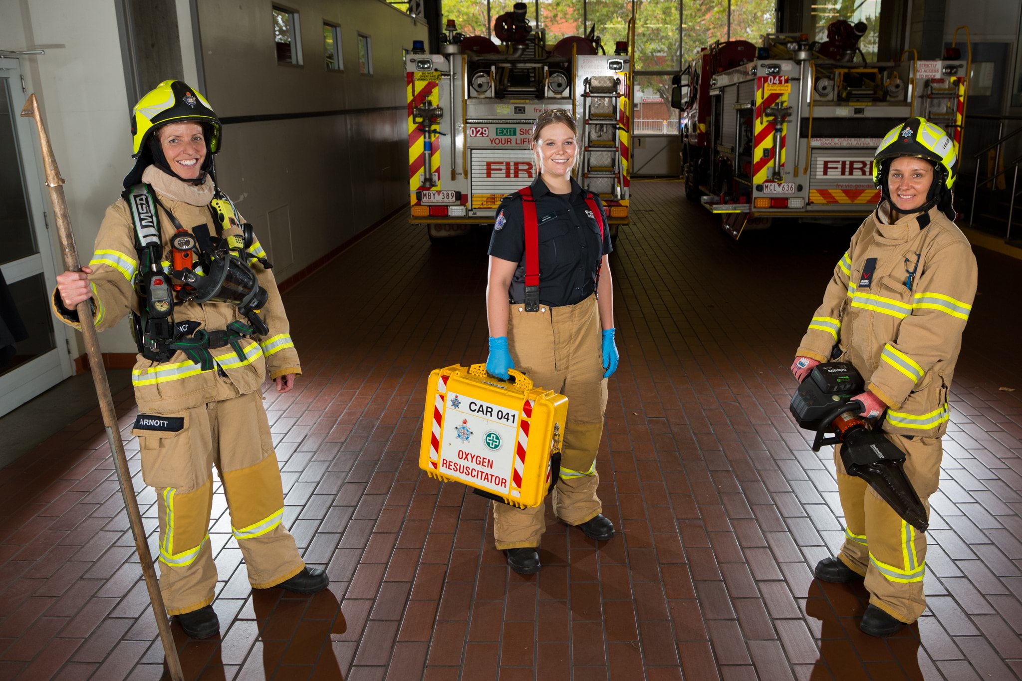 Photo of Fire Rescue Victoria Central District firefighters Emily Arnott, Ebony Lyons and Amanda Lawless. The three firefighters are dressed in their yellow uniforms and are holding specialist gear. In the background are two fire trucks. 